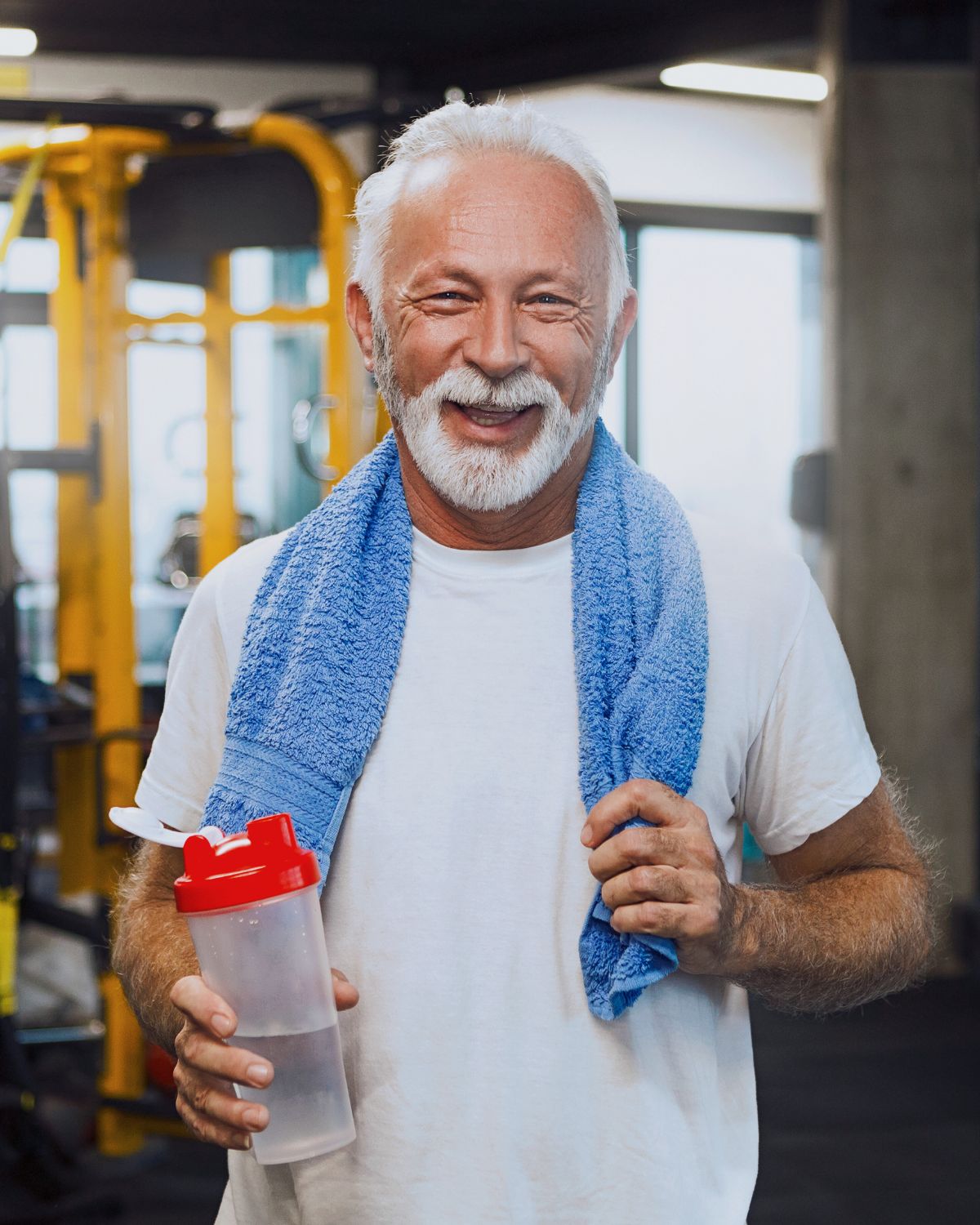 Äldre man på ett gym som tittar in i kameran med ett leende, med en blå handduk runt halsen och en vattenflaska i handen efter ett träningspass.